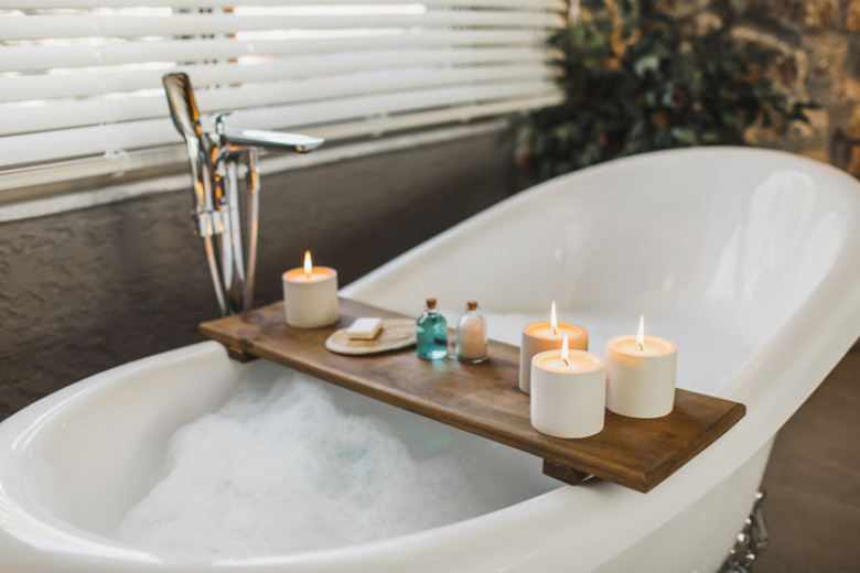 Wooden tray on bath tub with burning candles, bath salt and shampoo in bottles, loofah sponge and soap