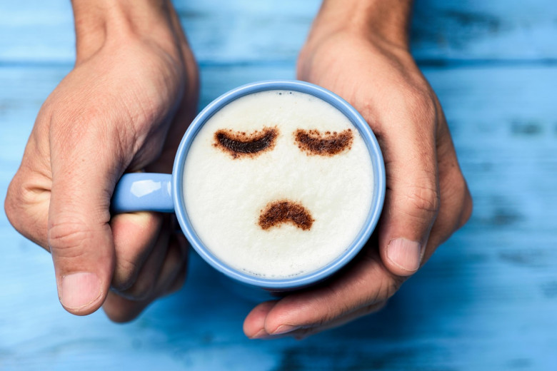 high-angle shot of a young caucasian man with a blue cup of cappuccino with a sad face drawn with cocoa powder on the milk foam, on a blue rustic tabl