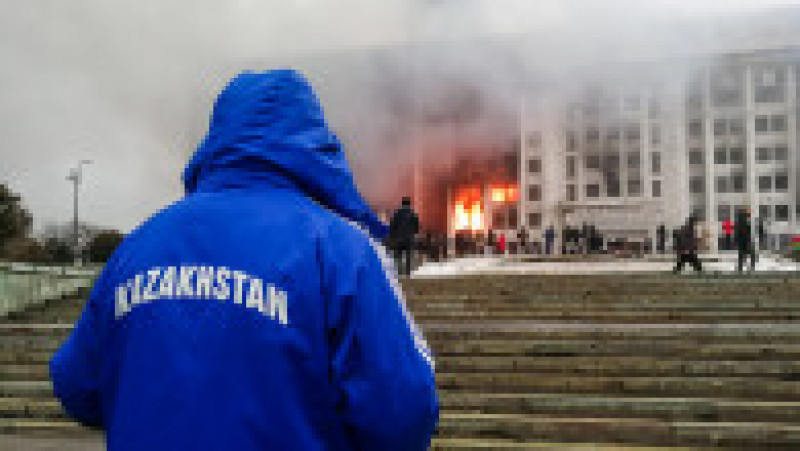 Un bărbat care poartă un trening pe care scrie „Kazahstan” se uită la clădirea în flăcări a primăriei din Almatî. Foto: Profimedia Images | Poza 17 din 24