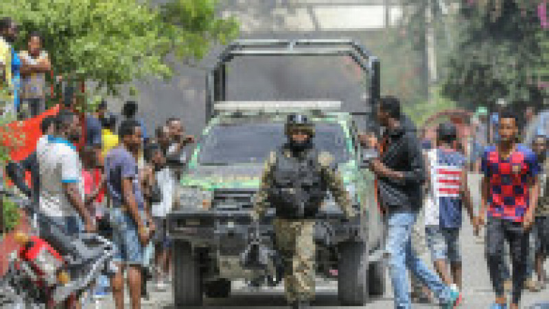 Oamenii se uită într-o mașină de poliție care trece pe stradă, în timp ce secția Petionville, unde sunt reținuți suspecții în cazul uciderii președintelui Jovenel Moise, este înconjurată. Sursa foto: Profimedia Images | Poza 10 din 26