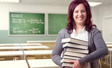Portrait Of Cheerful Teacher Holding Stacked Books In Classroom