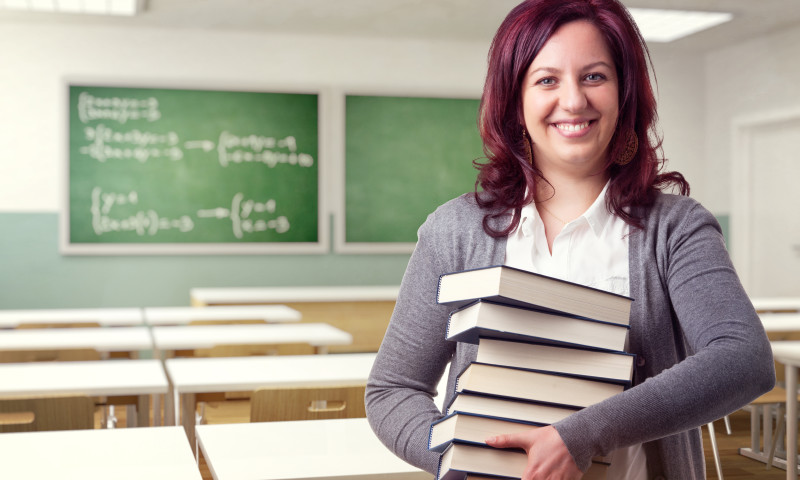 Portrait Of Cheerful Teacher Holding Stacked Books In Classroom